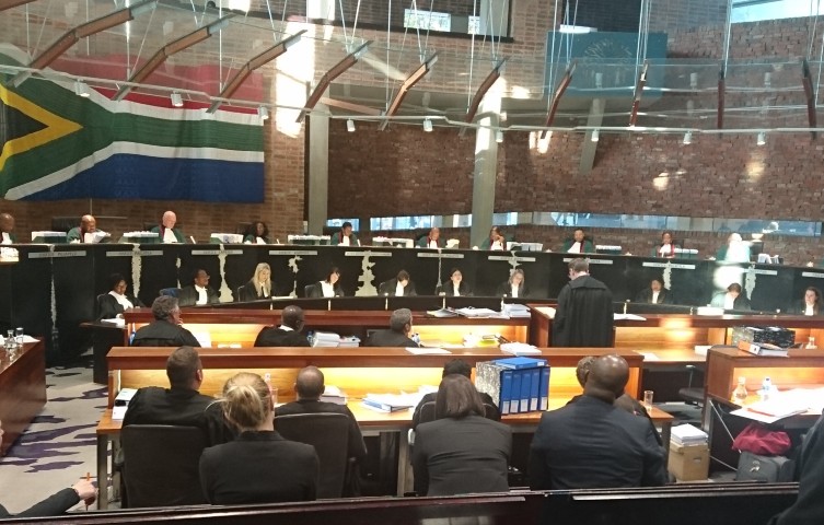 Judges and officials seated around a curved wooden bench in a formal chamber, with a large flag on the wall behind them.