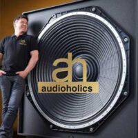 AH-SUB - Municipal Debt Specialist Man in a black polo leaning against a large circular speaker with the Audioholics logo on the cone.